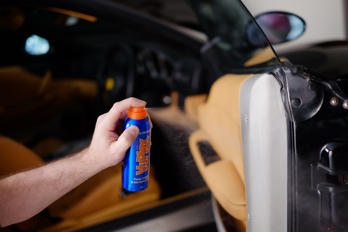 Hand holding a VuPlex bottle, applying plastic cleaner and polish in a car interior.
