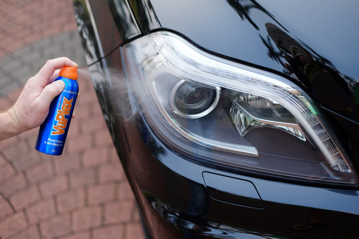 Hand holding VuPlex spray bottle applying cleaner to car headlight for effective plastic maintenance.
